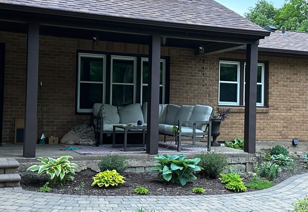 Photo 3) paved walk way, green plants in front of a porch. the porch is open with a brown post, on the porch is a small table and an l-shaped outdoor couch with cream colored cushions. 