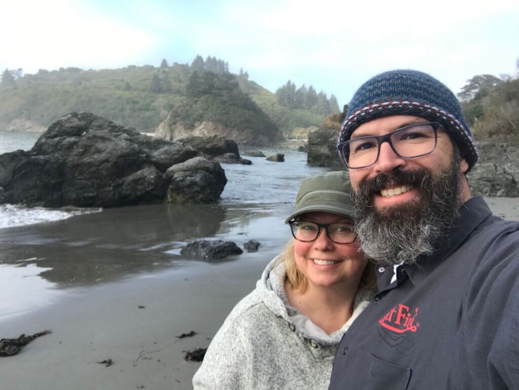 smiling couple on the CA coast, rocks, water, and pine trees in the background